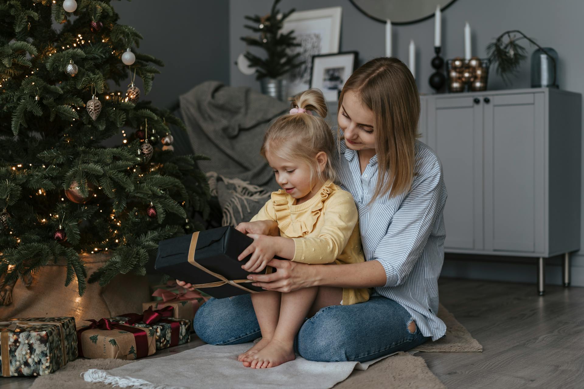 A toddler sits on her mothers lap looking at christmas presents.