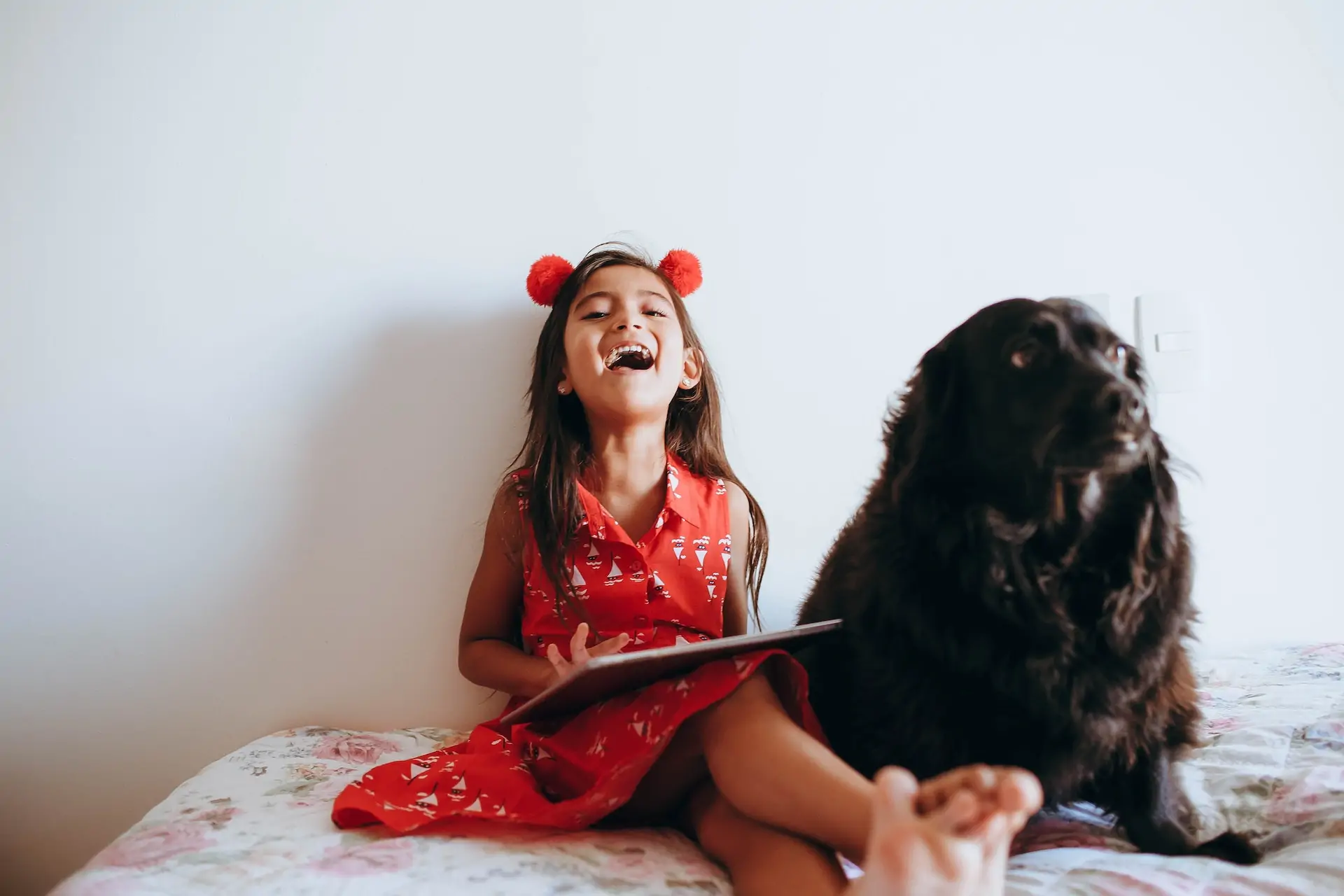 A girl sits on her bed with a book in her hand, laughing as her puppy sits beside her.